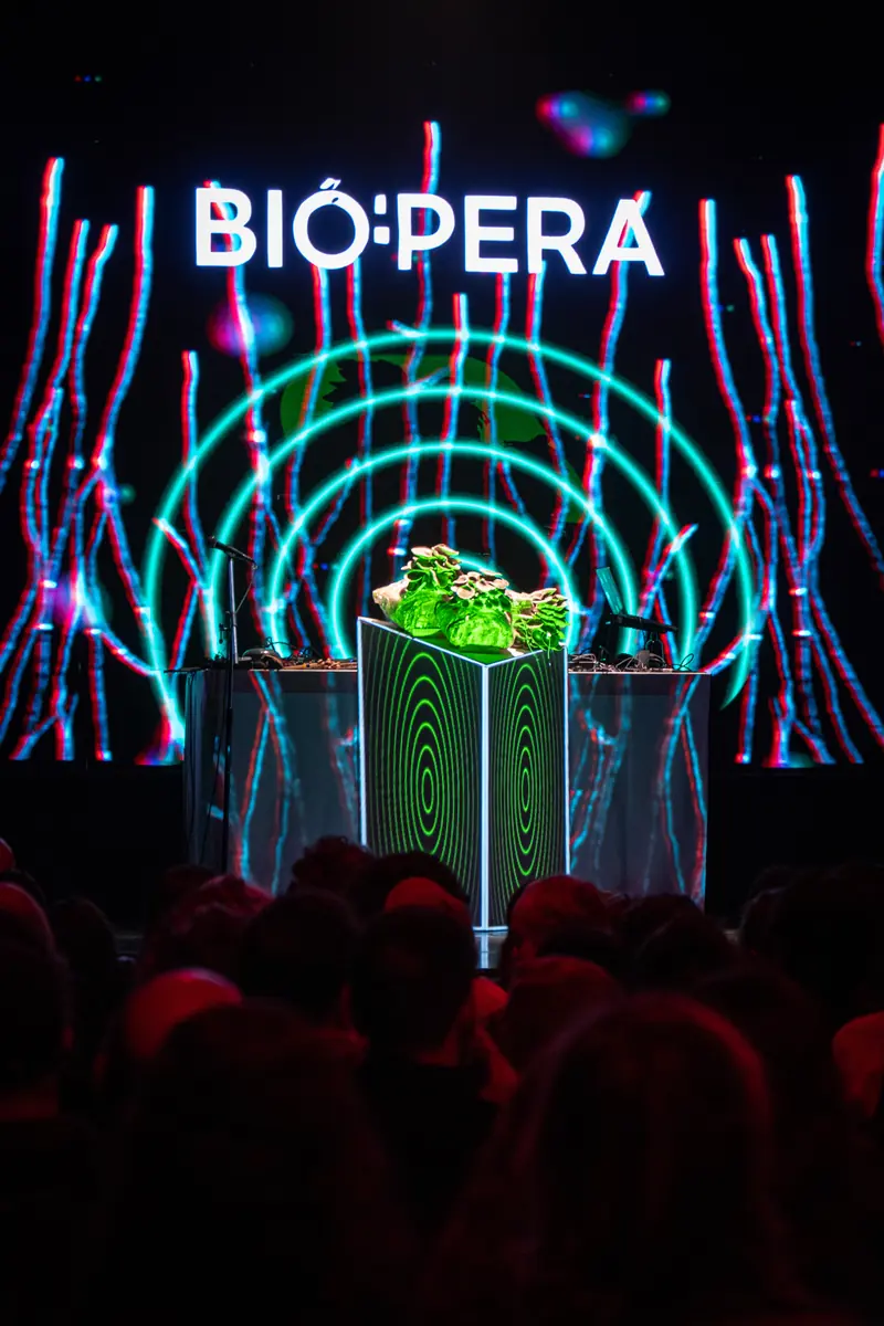 El logotipo BIÓ:PERA proyectado sobre la pantalla, con anillos turquesa, y debajo la mesa con el cultivo de hongos frente al público en penumbra