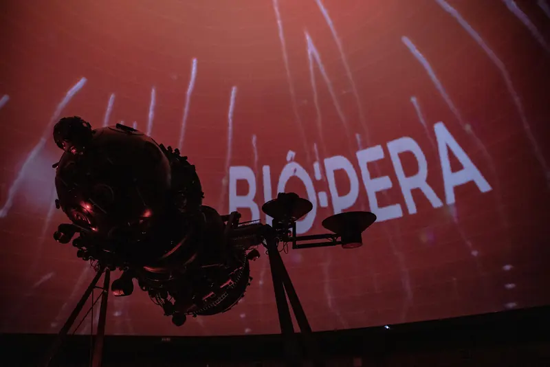 El proyector estelar del planetario, en silueta, contra el domo rojo con el logotipo BIÓ:PERA proyectado